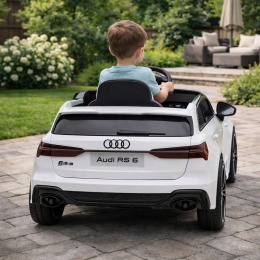 Dziecko jadące białym autem elektrycznym Audi RS6 na podwórku, widok od tyłu, zdjęcie produktowe w ogrodzie.
