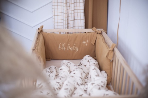Bawełniany ochraniacz do łóżeczka w kolorze karmelowym z haftem Oh Baby i dekoracyjnymi wiązaniami.